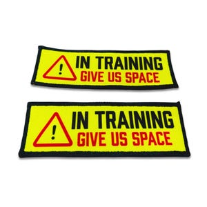 Op de afbeelding: Twee rechthoekige patches met een felgele achtergrond en een zwarte rand. Elke patch heeft links een rode driehoek met een uitroepteken, gevolgd door de zwarte tekst IN TRAINING en de rode tekst GIVE US SPACE.