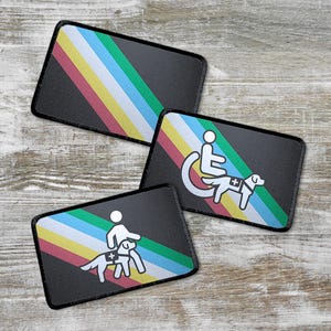 Op de afbeelding: Drie zwarte patches met een regenboogstreepontwerp. Elke patch toont een wit silhouet van een persoon met een hulphond. Eén patch toont een persoon in een rolstoel, een andere toont een persoon die loopt en de derde toont een persoon die staat.