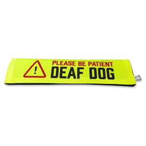 Op de afbeelding: Een rechthoekige neongele band met een zwarte rand. Het bevat een rode driehoek met een uitroepteken, gevolgd door de rode tekst PLEASE BE PATIENT en de dikgedrukte zwarte tekst DEAF DOG. Een klein wit stoffen label zit aan de zijkant.