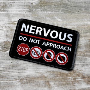 Könnte beinhalten: Schwarz-weißes Patch mit roten Akzenten. Das Patch sagt "NERVOUS DO NOT APPROACH" mit einem Stoppschild, einem durchgestrichenen Auge, einer durchgestrichenen Hand und einer durchgestrichenen Sprechblase.