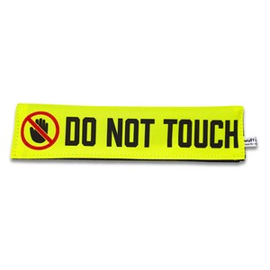 Könnte beinhalten: Ein rechteckiges, neongelbes Stoffband mit dem fetten schwarzen Text DO NOT TOUCH und einem roten kreisförmigen Symbol mit einer schwarzen Hand darin. Ein kleines weißes Stoffetikett mit schwarzem Text ist an der rechten Seite befestigt.
