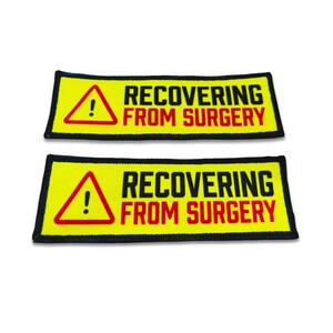 Op de afbeelding: Twee rechthoekige patches met een felgele achtergrond en een zwarte rand. Elke patch heeft links een rode driehoek met een zwart uitroepteken en rechts de zwarte en rode tekst RECOVERING FROM SURGERY in hoofdletters gedrukt.
