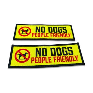 Op de afbeelding: Twee rechthoekige geborduurde patches met een felgele achtergrond en zwarte randen. Elke patch bevat een rode cirkel met een diagonale lijn over een zwart hondensilhouet, gevolgd door de zwarte tekst NO DOGS en rode tekst PEOPLE FRIENDLY.