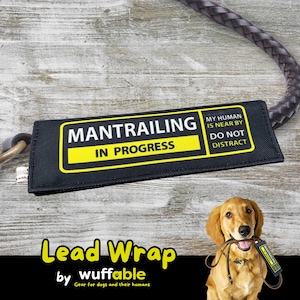 Könnte beinhalten: Schwarzes Hundeleinen-Etikett mit dem Text "MANTRAILING IN PROGRESS" in Weiß und Gelb. Auf dem Etikett steht auch "MY HUMAN IS NEAR BY DO NOT DISTRACT". Ein Golden Retriever hält ein passendes Etikett. Die Wörter "Lead Wrap" und "wuffable" sind ebenfalls sichtbar.