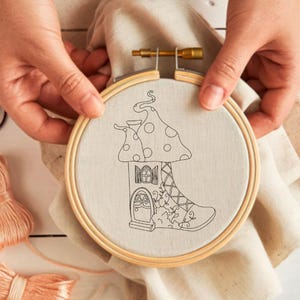 Puede incluir: Bastidor de bordado con un diseño de casa hongo. El diseño es un contorno negro sobre una tela beige. El bastidor es sostenido por manos, con madejas de hilo en tonos melocotón, verde y burdeos cerca. El bastidor de madera tiene 15 cm de diámetro.