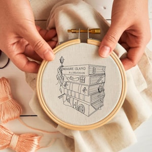 Puede incluir: Un bastidor de bordado con una tela de lino con una ilustración en blanco y negro de una pila de libros con el texto "TREASURE ISLAND R.L. STEVENSON". El bastidor es sostenido por manos, con carretes de hilo en el fondo.