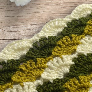 May include: Close-up of a crocheted item with a shell stitch pattern. The yarn colors include cream, olive green, and a golden yellow. The crochet work is displayed on a wooden surface.