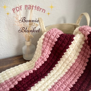 Könnte beinhalten: Eine gehäkelte Decke mit Streifen aus weißem, rosa und burgunderfarbenem Garn. Die Decke ist gefaltet und in einem geflochtenen Korb gelegt. Der Text "PDF Pattern Bonnie Blanket" ist im Bild sichtbar.