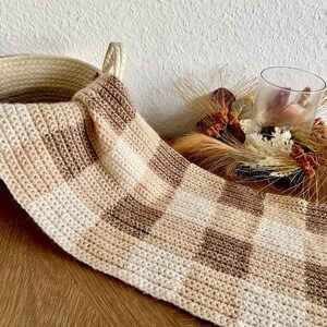 May include: A crocheted throw blanket with a checkered pattern in beige and brown tones. The blanket is draped over a woven basket and a wooden surface.