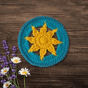 May include: A crocheted sun design with a yellow center and blue outer ring. The sun is crocheted in a circular shape and has a textured appearance.