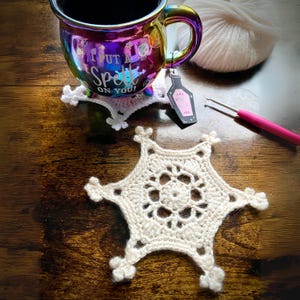May include: A white crocheted snowflake coaster with a detailed design. The coaster is sitting on a wooden table next to a mug with the text "I put a spell on you!" and a small bottle charm.
