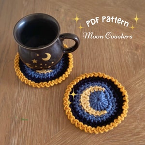 Peut inclure: Deux dessous-de-verre en crochet en forme de lune, dans des tons de bleu et de jaune, avec une bordure dorée. Les dessous-de-verre sont posés sur une surface en bois. Le texte "PDF Pattern Moon Coasters" est visible sur l'image.