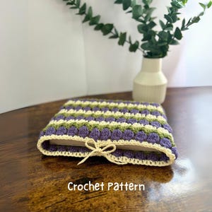 May include: A crocheted book cover with a striped pattern in shades of purple, green, and cream. The cover has a cream-coloured ribbon tie. The text "Crochet Pattern" is visible below the cover.