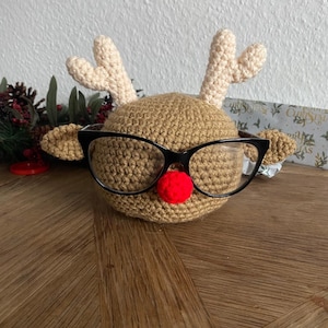 Peut inclure: Une tête de renne au crochet avec des lunettes noires et un nez rouge. Le renne a des bois et des oreilles beiges. Il est posé sur une surface en bois.