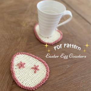 May include: Two crocheted Easter egg coasters in white with pink trim and pink flower accents. The coasters are on a wooden surface with a white mug on one of the coasters. The text "PDF Pattern Easter Egg Coasters" is on the image.
