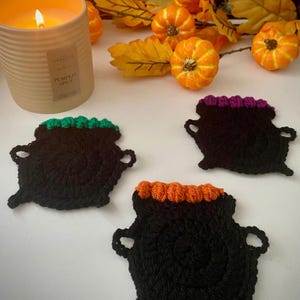 Puede incluir: Tres posavasos negros de ganchillo con forma de caldero con la parte superior verde, naranja y morada. Una vela encendida y pequeñas calabazas en el fondo sugieren un tema de Halloween o otoño. La vela tiene la inscripción "Pumpkin Spice".