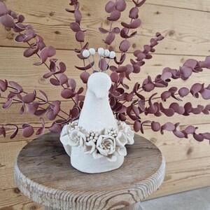Peut inclure: Une figurine d'oiseau en céramique blanche avec une couronne florale et une petite couronne de sphères blanches. L'oiseau est posé sur un petit tabouret rond en bois. Derrière l'oiseau, des branches de feuilles d'eucalyptus bordeaux.