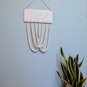Peut inclure: Une suspension murale en céramique blanche avec un motif floral délicat et une série de perles en bois qui pendent. Les perles sont disposées en cascade, créant une décoration murale visuellement attrayante et texturée.