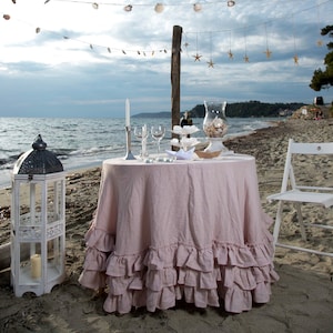 Puede incluir: Una mesa romántica preparada en la playa con un mantel rosa con volantes, sillas blancas y una guirnalda de conchas marinas por encima. La mesa está decorada con velas, vasos y un plato de postres.