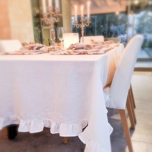 May include: White linen tablecloth with a ruffled edge, set on a dining table with white chairs. The table is decorated with place settings and a candelabra with lit candles.