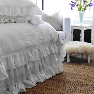 May include: White ruffled bed linens with a white throw pillow and a white footstool in a bedroom with a brown woven rug. A vase of purple irises sits on a white table.
