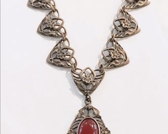 May include: A silver-toned necklace with an oval, red gemstone pendant. The necklace features a series of floral-shaped links and a decorative pendant setting. The pendant is approximately 1.5 inches long.