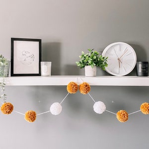 May include: A white shelf with a black framed print, a small potted plant, a white candle, a white clock, and a black candle. A garland of white and brown pom-poms hangs below the shelf.
