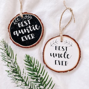 Könnte beinhalten: Zwei runde Holzscheiben-Ornamente mit Bindfadenaufhängern. Ein Ornament ist schwarz mit weißem Text, der "Best auntie ever" lautet. Das andere Ornament ist weiß mit schwarzem Text, der "Best uncle ever" lautet.