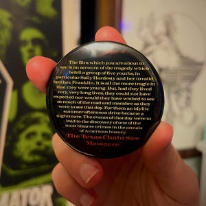 Puede incluir: Un botón negro con texto de la película "The Texas Chain Saw Massacre". El texto amarillo describe la trama de la película, mencionando a Sally Hardesty y Franklin. El botón es sostenido por una persona.