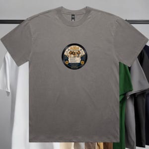 May include: A charcoal grey short-sleeve t-shirt featuring a circular graphic on the chest. The graphic depicts a vinyl record label with the text ABBA, The Album, and a list of song titles, centred on the shirt with a crew neckline and hem.