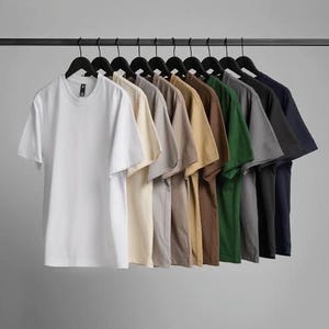 K&ouml;nnte beinhalten: Eine Reihe von einfarbigen T-Shirts mit kurzen &Auml;rmeln in verschiedenen Farben, darunter Wei&szlig;, Beige, Grau, Braun, Gr&uuml;n und Marineblau, h&auml;ngen an schwarzen Kleiderb&uuml;geln vor grauem Hintergrund. Die Shirts sind aus weichem Material.