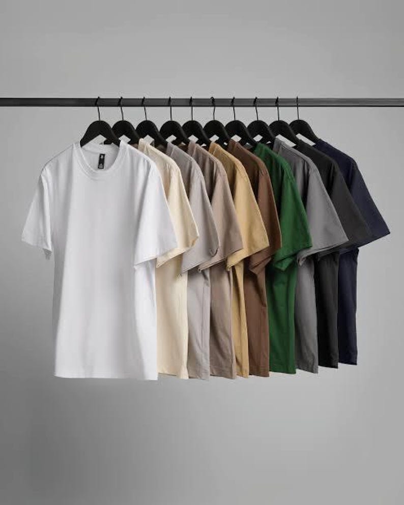 K&ouml;nnte beinhalten: Eine Reihe von einfarbigen T-Shirts mit kurzen &Auml;rmeln in verschiedenen Farben, darunter Wei&szlig;, Beige, Grau, Braun, Gr&uuml;n und Marineblau, h&auml;ngen an schwarzen Kleiderb&uuml;geln vor grauem Hintergrund. Die Shirts sind aus weichem Material.