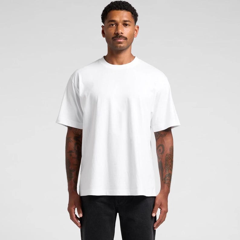 K&ouml;nnte beinhalten: Ein schlichtes, wei&szlig;es T-Shirt mit kurzen &Auml;rmeln. Das Shirt hat einen Rundhalsausschnitt und eine lockere Passform. Das Model tr&auml;gt eine schwarze Hose. Der Hintergrund ist einfarbig hellgrau.