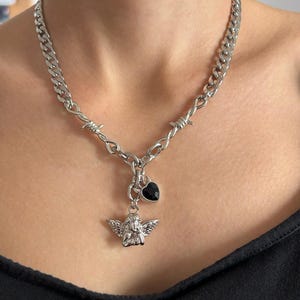 Puede incluir: Collar plateado con diseño de alambre de púas, un pequeño colgante de ángel y un dije de corazón negro. El collar tiene un estilo de cadena y se usa alrededor del cuello.