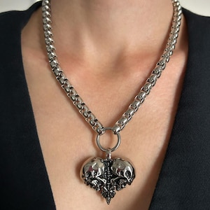 May include: Silver-toned necklace featuring a heart-shaped pendant with skull detailing. The pendant is suspended from a thick, rounded chain. The necklace is worn against a dark background.