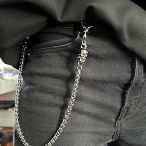 May include: A silver chain with a skull pendant attached to black jeans. The chain is made of small, square links and hangs from the pocket. The skull charm is detailed, adding a gothic element to the accessory. The jeans are dark grey.