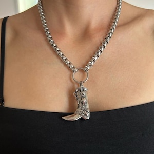 May include: Silver-toned necklace featuring a detailed cowboy boot pendant. The pendant showcases intricate designs, including skulls, and hangs from a sturdy chain. The necklace is a bold accessory, suitable for adding a touch of Western flair.