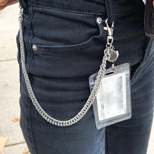 K&ouml;nnte beinhalten: Eine silberne Kette mit einem durchsichtigen Plastikkartenhalter, der an einem silbernen Clip befestigt ist. Die Kette h&auml;ngt an einer schwarzen Jeans.