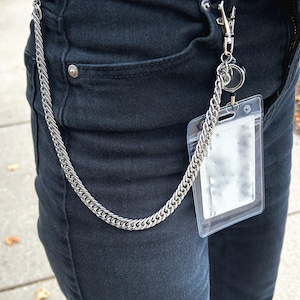 May include: A silver chain with a clear plastic ID card holder attached to it. The chain is hanging from a person's belt loop.