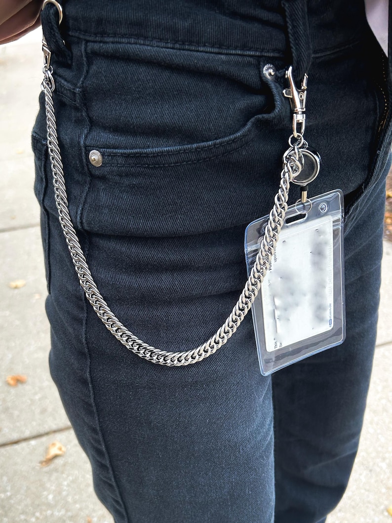 K&ouml;nnte beinhalten: Eine silberne Kette mit einem durchsichtigen Plastik-Ausweishalter daran. Die Kette h&auml;ngt an einer G&uuml;rtelschlaufe.