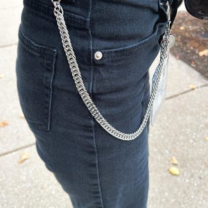 K&ouml;nnte beinhalten: Eine silberne Kette mit einem Verschluss, die an einer schwarzen Jeans befestigt ist.