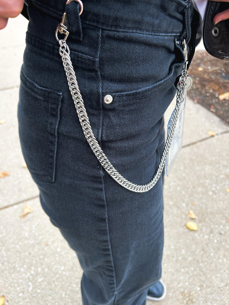K&ouml;nnte beinhalten: Eine silberne Kette mit einem Verschluss, die an einer schwarzen Jeans befestigt ist.
