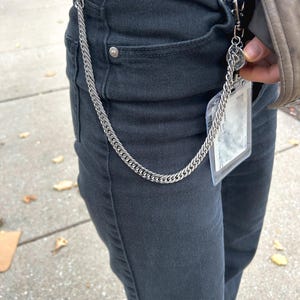 K&ouml;nnte beinhalten: Eine silberne Kette mit einem Verschluss, die an einer schwarzen Jeans befestigt ist. Die Kette h&auml;ngt an der Vorderseite der Jeans herunter.