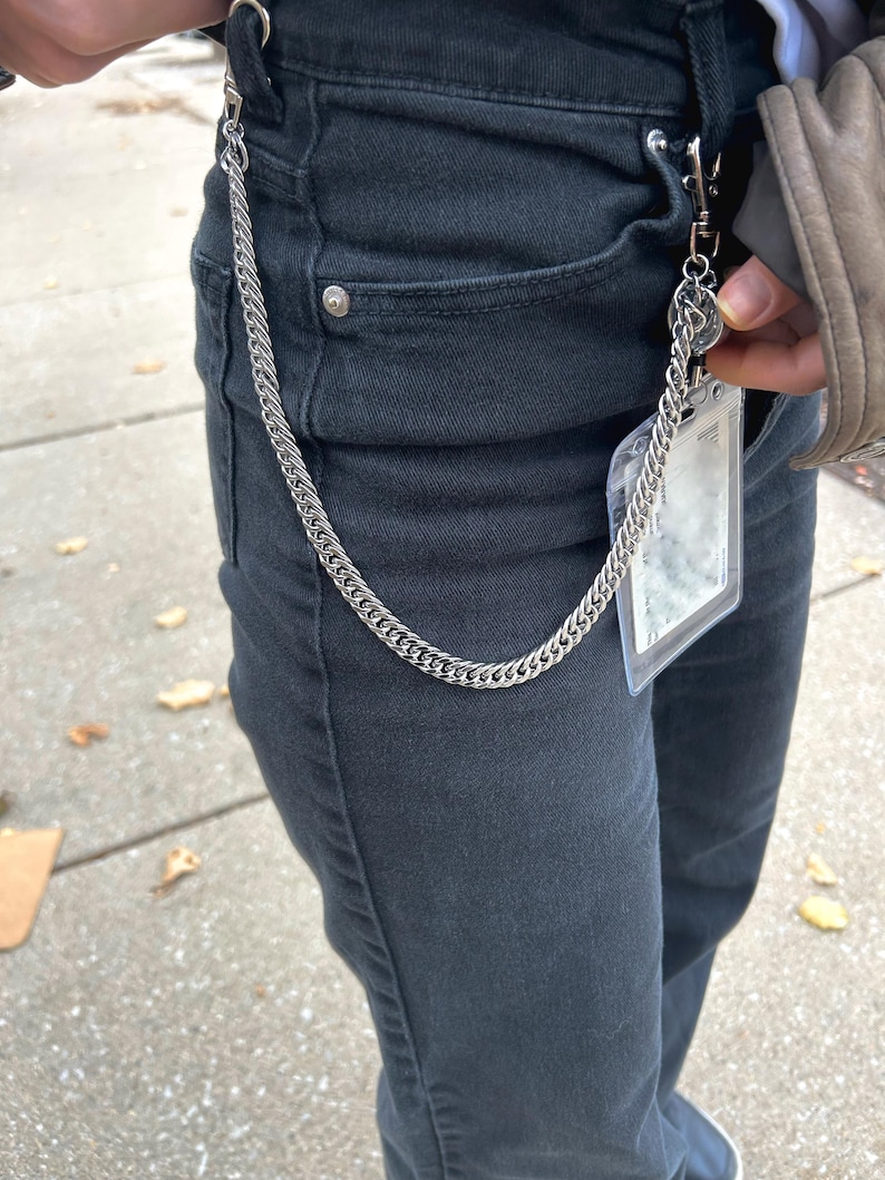 K&ouml;nnte beinhalten: Eine silberne Kette mit einem Verschluss, die an einer schwarzen Jeans befestigt ist. Die Kette h&auml;ngt an der Vorderseite der Jeans herunter.