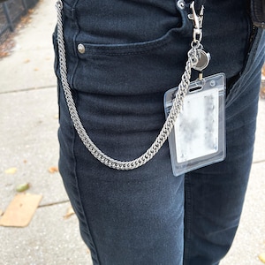 K&ouml;nnte beinhalten: Eine silberne Kette mit einem silbernen Verschluss, der an einem durchsichtigen Plastik-Ausweishalter befestigt ist. Die Kette h&auml;ngt an der vorderen Tasche einer schwarzen Jeans.