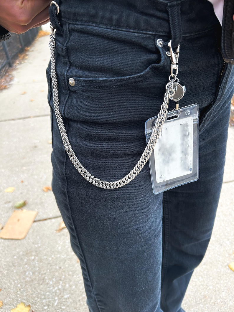 K&ouml;nnte beinhalten: Eine silberne Kette mit einem silbernen Verschluss, der an einem durchsichtigen Plastik-Ausweishalter befestigt ist. Die Kette h&auml;ngt an der vorderen Tasche einer schwarzen Jeans.