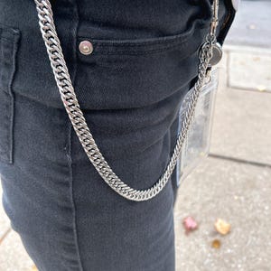 K&ouml;nnte beinhalten: Eine silberne Kette mit einem durchsichtigen Plastik-Ausweishalter. Die Kette h&auml;ngt an der vorderen Tasche einer schwarzen Jeans.