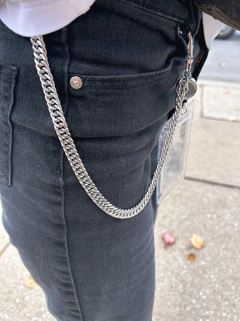 K&ouml;nnte beinhalten: Eine silberne Kette mit einem durchsichtigen Plastik-Ausweishalter. Die Kette h&auml;ngt an der vorderen Tasche einer schwarzen Jeans.