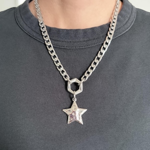 Puede incluir: Un collar de tono plateado con una cadena gruesa de eslabones tipo barbada y un conector hexagonal. Un colgante de estrella de cinco puntas con una superficie martillada y texturizada cuelga del centro. El metal tiene un acabado pulido.
