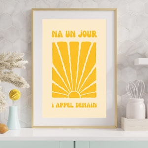 Peut inclure: Une affiche encadrée avec un cadre doré. L'impression présente un motif de soleil jaune sur un fond crème, avec le texte "NA UN JOUR" et "! APPEL DEMAIN" en jaune. L'œuvre est exposée sur un comptoir blanc.